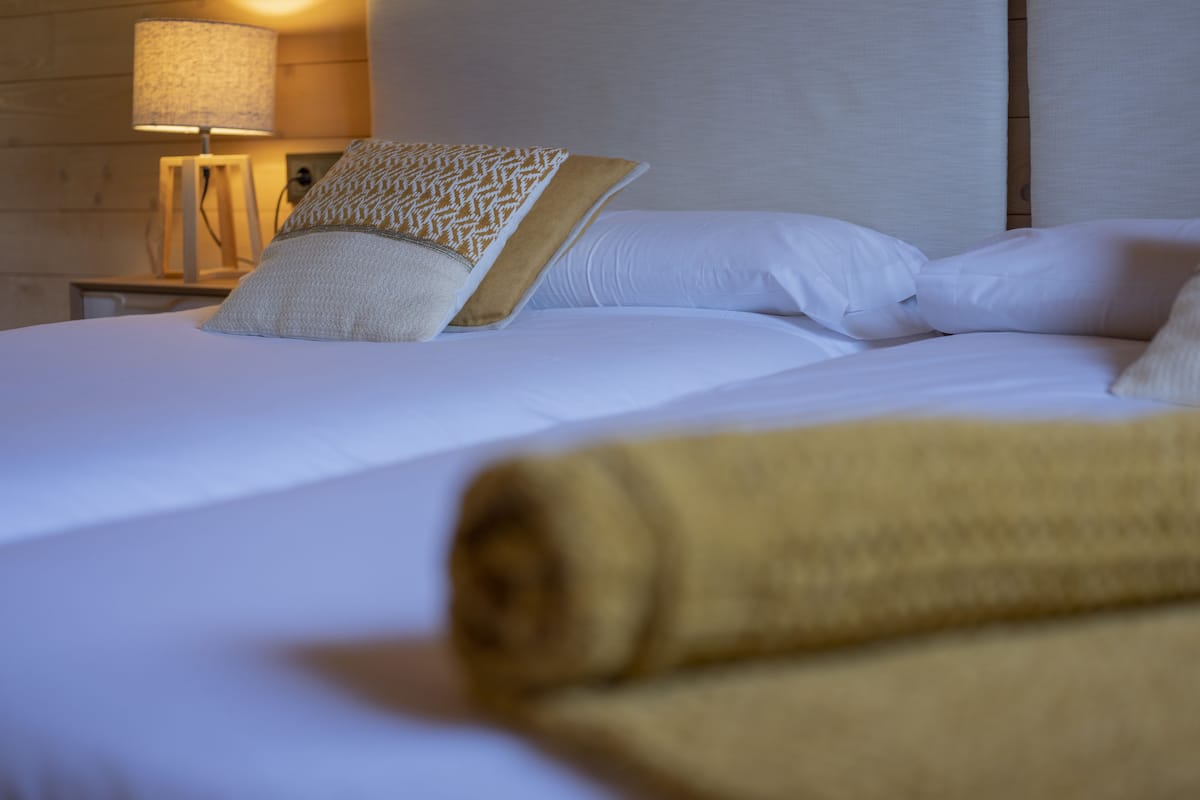 Una cama bien hecha con sábanas blancas, una toalla amarilla enrollada, un cojín estampado y una lámpara en una mesita de noche.