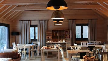 A spacious dining area with wooden tables and chairs, a central buffet table with food, and a large window with curtains.