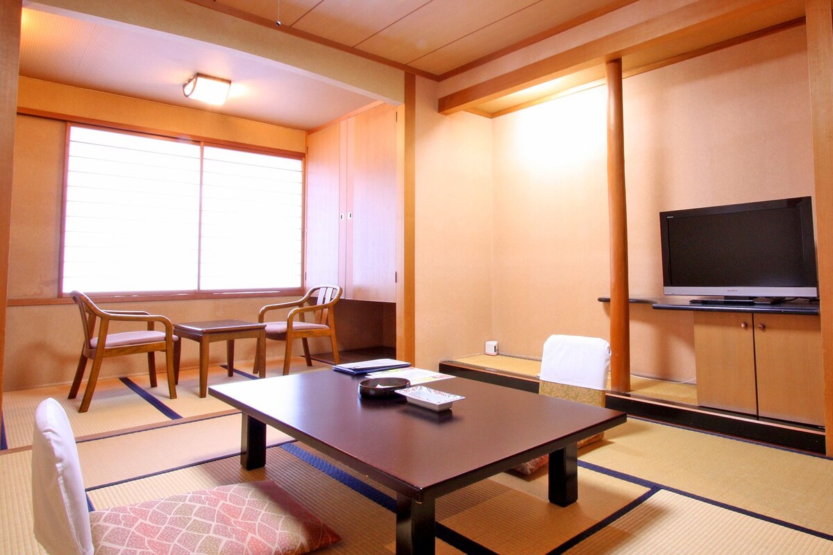Une pièce de style japonais traditionnel, avec un sol recouvert de tatamis, une table basse, des chaises, un téléviseur et des portes coulissantes.