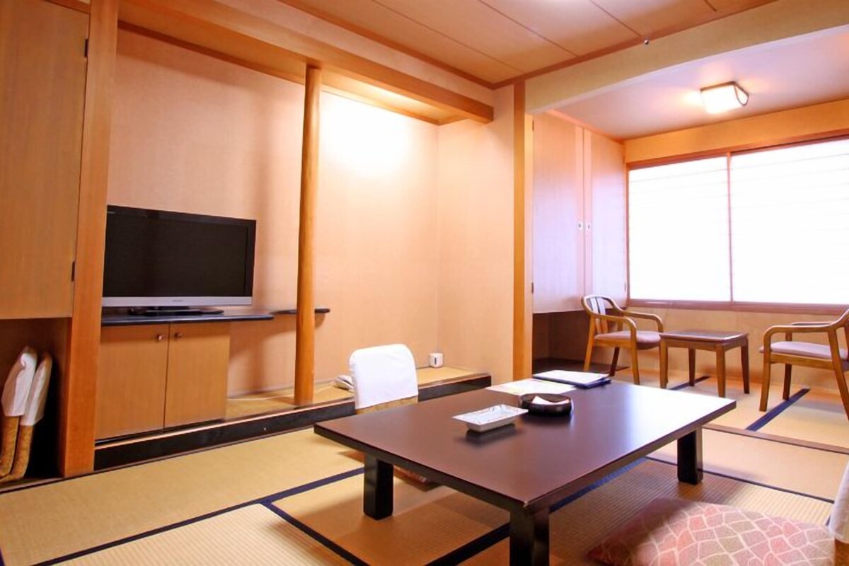 Una habitación tradicional japonesa con suelo de tatami, televisor, mesa con sillas y ventana con persianas.