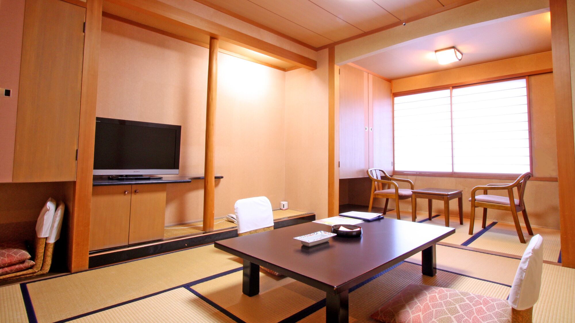 Una habitación japonesa tradicional con suelo de tatami, un televisor sobre un bajo pedestal, una mesa con cojín y una ventana con persianas.