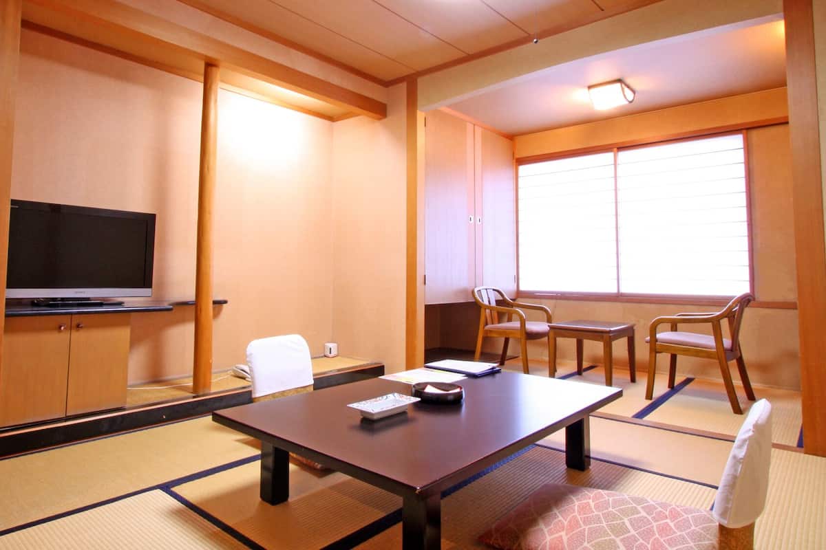 Habitació japonesa tradicional amb sòls de tatami, una televisió sobre un pedestal baix, una taula amb coixí i una finestra amb persianes.