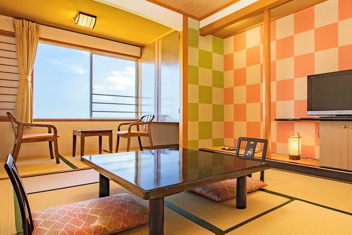 Habitació d'hotel amb sòl de tatami, una taula de dinar, cadires, un televisor i una finestra amb cortines.