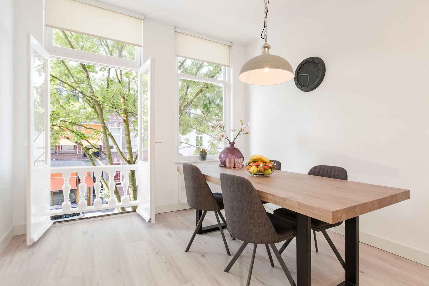 A dining area with a wooden table, four chairs, a hanging pendant light, a fruit bowl, and a clock on the wall.