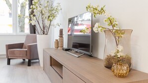 Moderni olohuone, jossa on televisio puisen tv-tason päällä, beigenvärinen nojatuoli ja valkoisia kukkia sisältävä maljakko.
