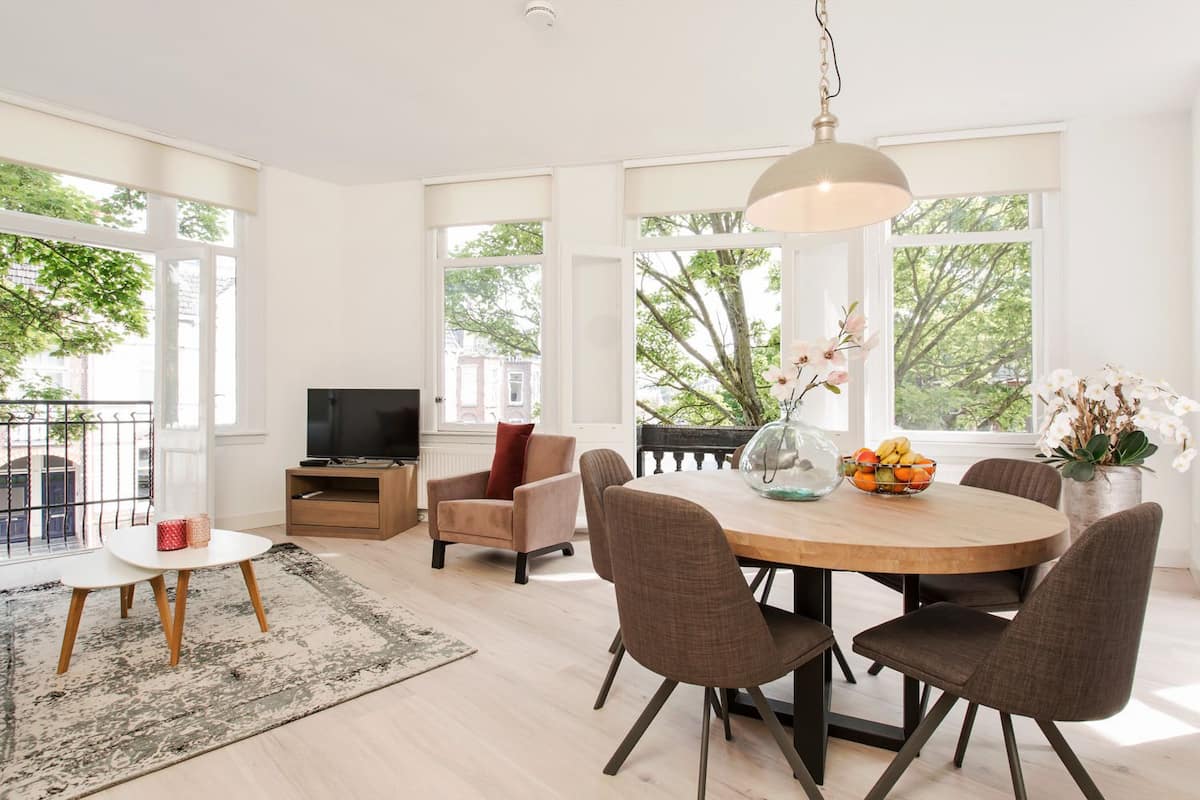 A modern living room with a dining area, large windows, and a view of trees.