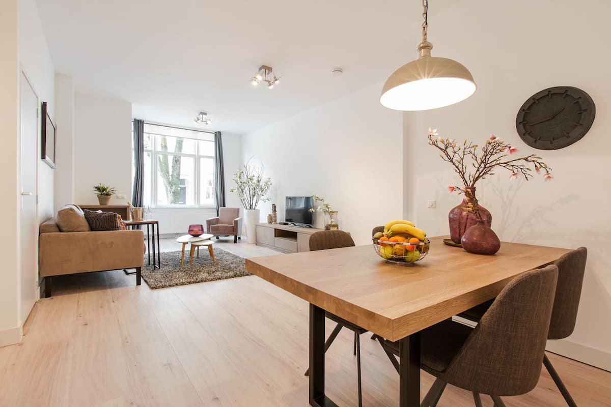 A modern living room with a wooden dining table, a sofa, a TV, and a large window.