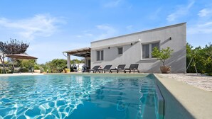 Un área moderna junto a la piscina, con piscina cristalina, tumbonas y un edificio con porche cubierto.