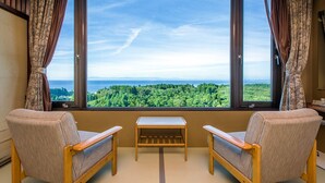 A room with two wooden chairs, a small table, and a view of a forest and coastline through large windows.