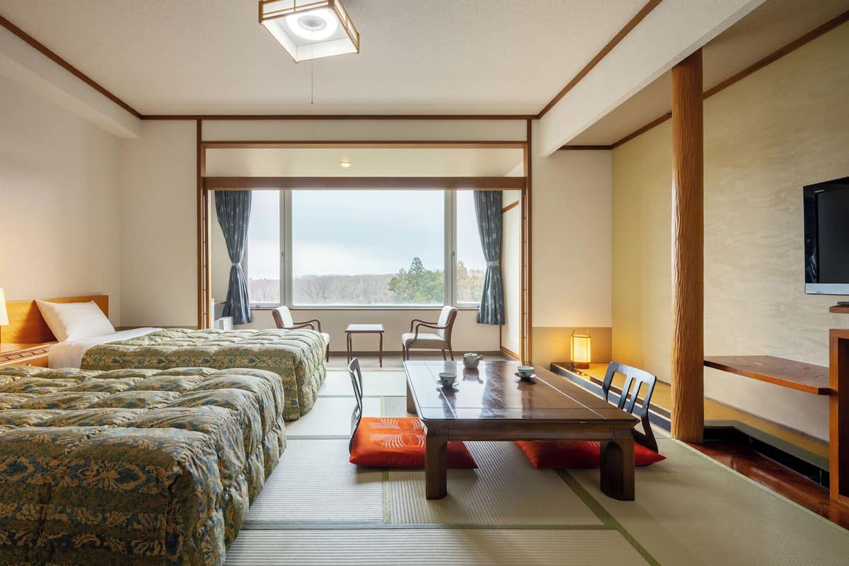 Ein Hotelzimmer mit zwei Betten, einem Esstisch, Stühlen, einem Fernseher und einem großen Fenster.