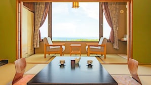 A room with a tatami mat floor, two wooden chairs, a table with a teapot and cups, and a view of the sea through large windows.