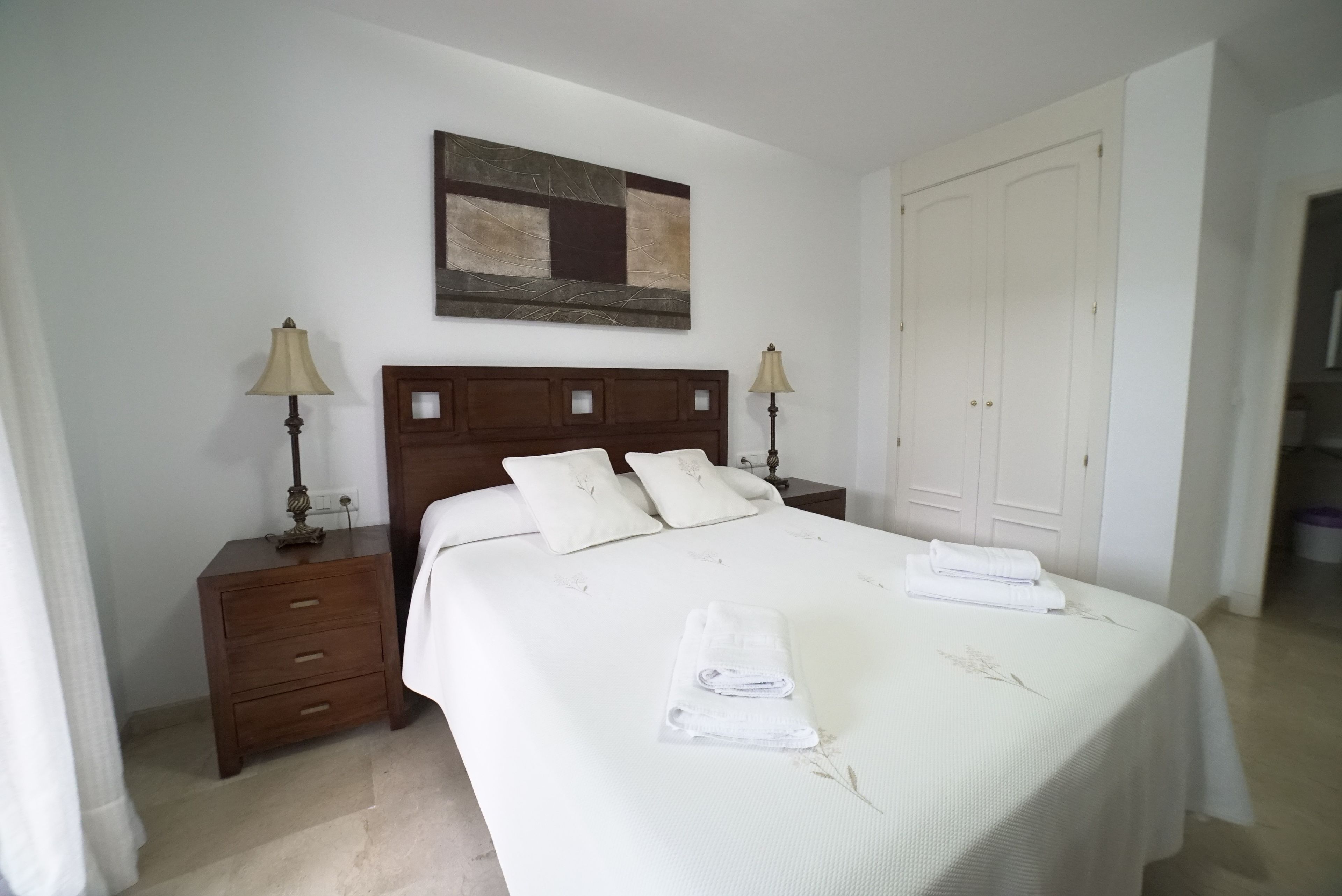 Una cama bien hecha con una colcha blanca y dos almohadas, una mesita de noche de madera con una lámpara, una obra de arte en la pared y un armario blanco.