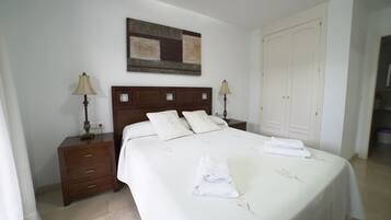 Una cama bien hecha con una colcha blanca y dos almohadas, una mesita de noche de madera con una lámpara, una obra de arte en la pared y un armario blanco.