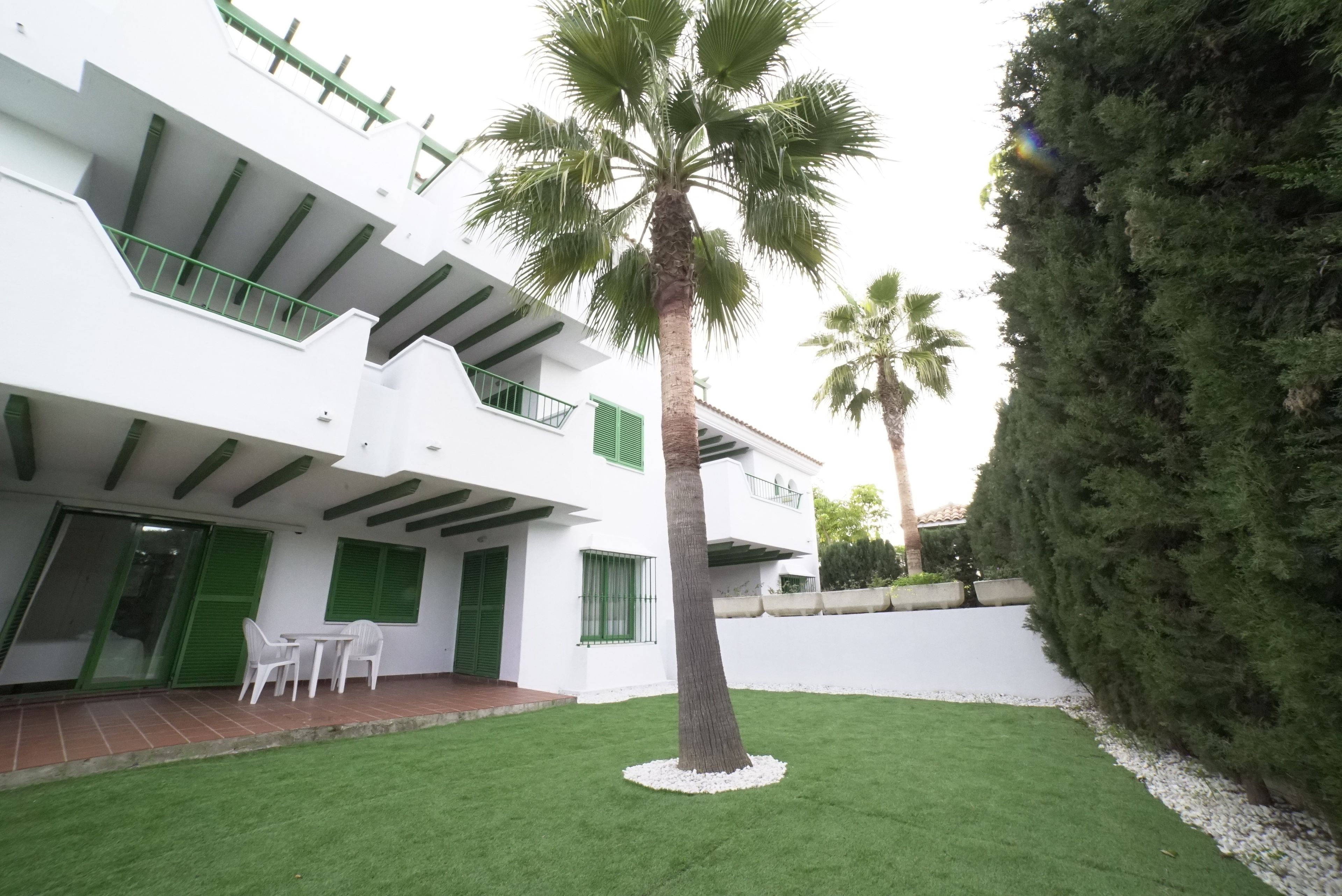 Un edificio moderno de dos pisos con techo plano, contraventanas verdes y un pequeño patio con una palmera y sillas de plástico blancas.