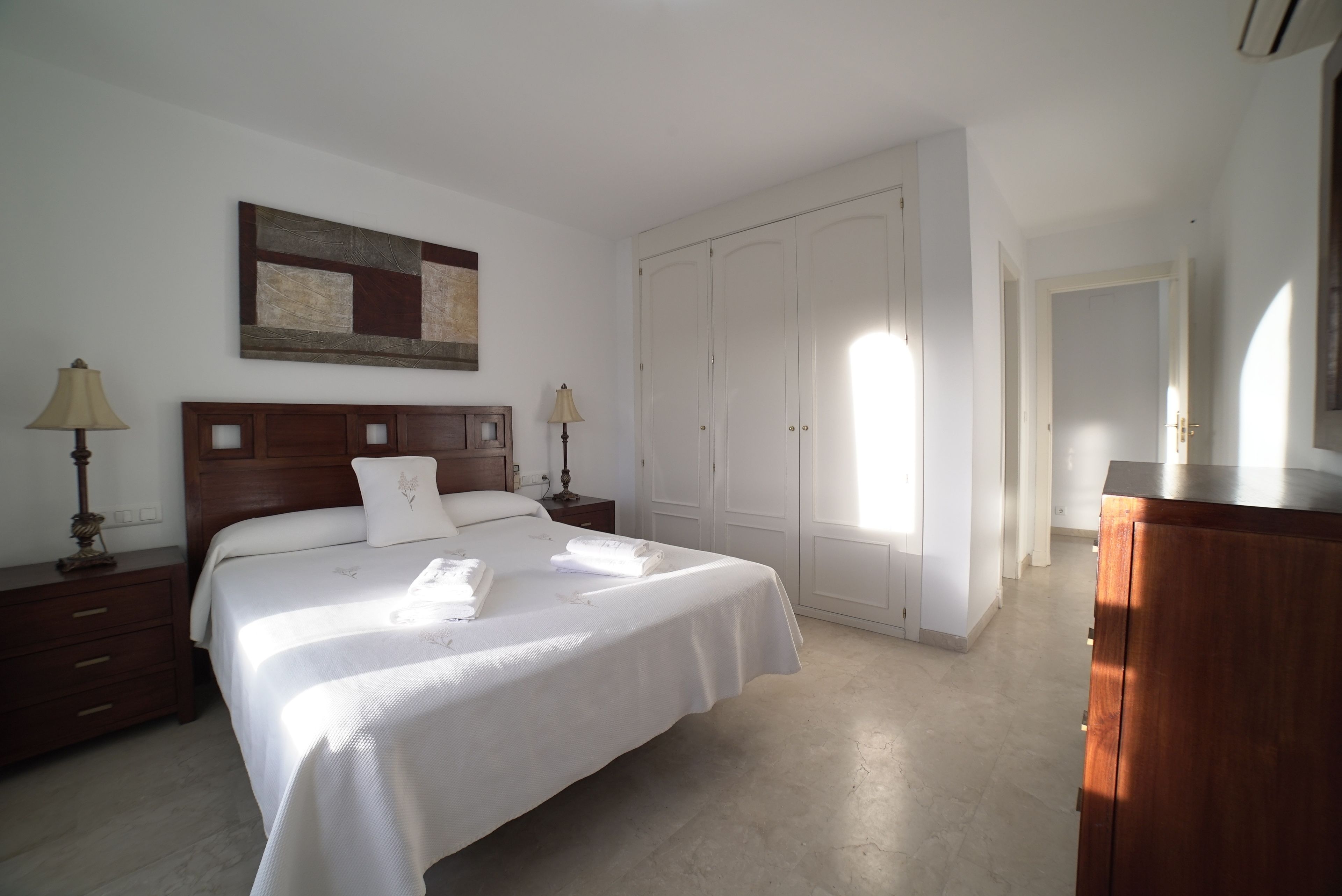 Un dormitorio con una cama grande, cabecera de madera, dos mesitas de noche con lámparas, un armario y un cuadro en la pared.