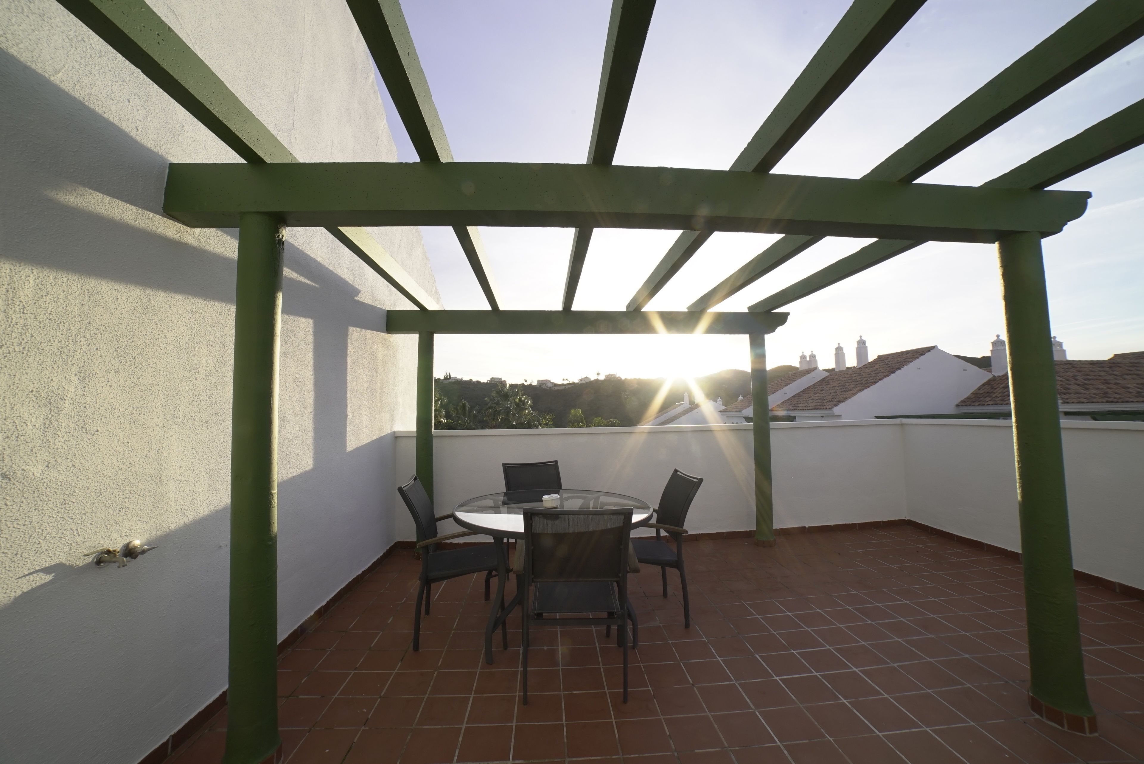 Eine überdachte Terrasse im Freien mit einem runden Tisch und Stühlen, einer Pergola und Blick auf Gebäude im Hintergrund.