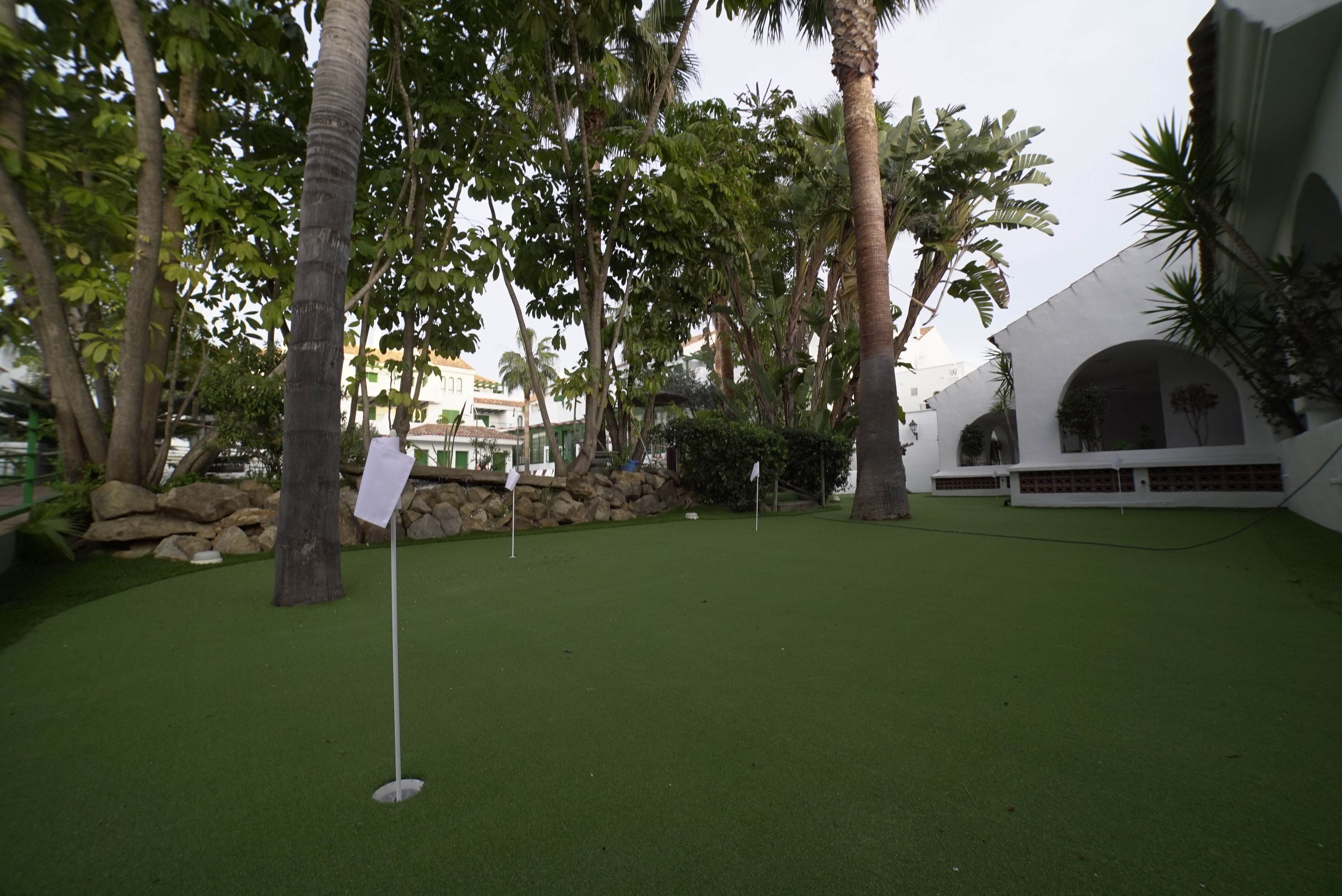 Een golfbaan met een putting green, omgeven door palmbomen en een wit gebouw met bogen.