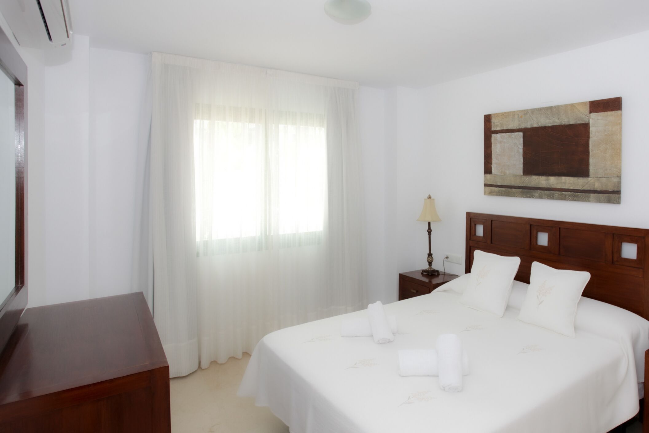 A hotel room with a bed, a nightstand, a lamp, a painting on the wall, and a window with curtains.