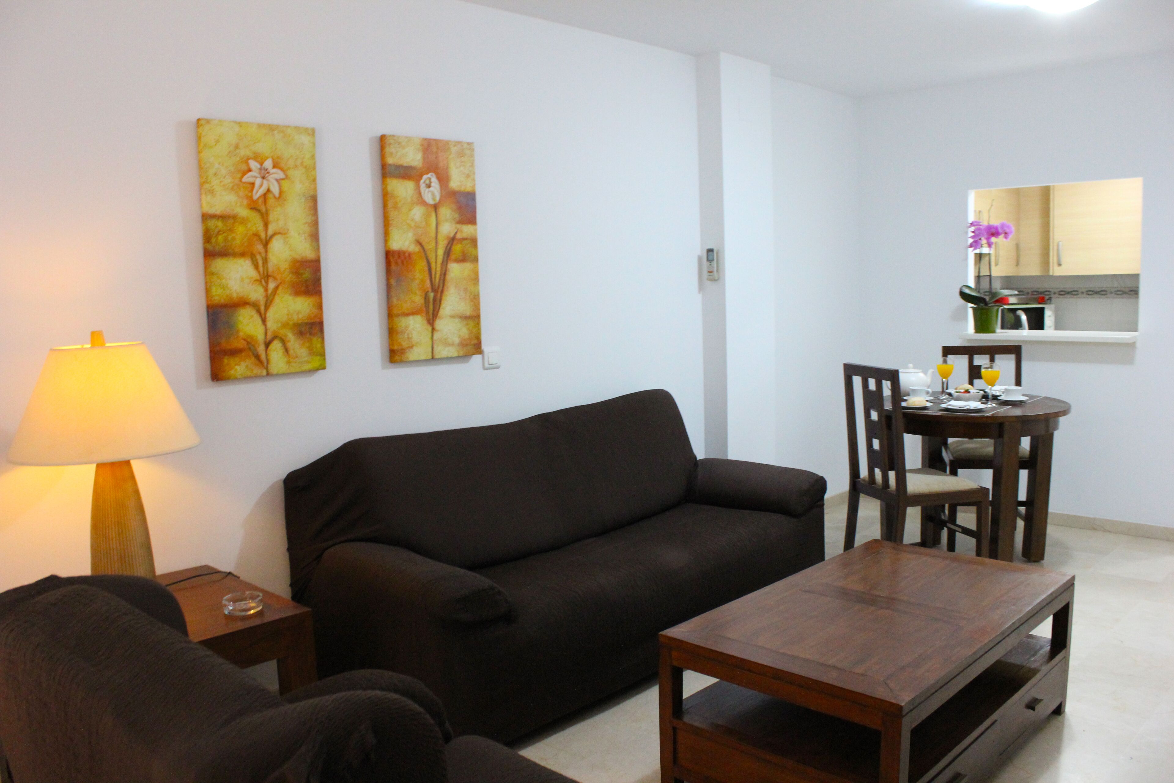 A living room with a sofa, a coffee table, and a dining table with chairs. There are two paintings on the wall and a lamp on a side table.