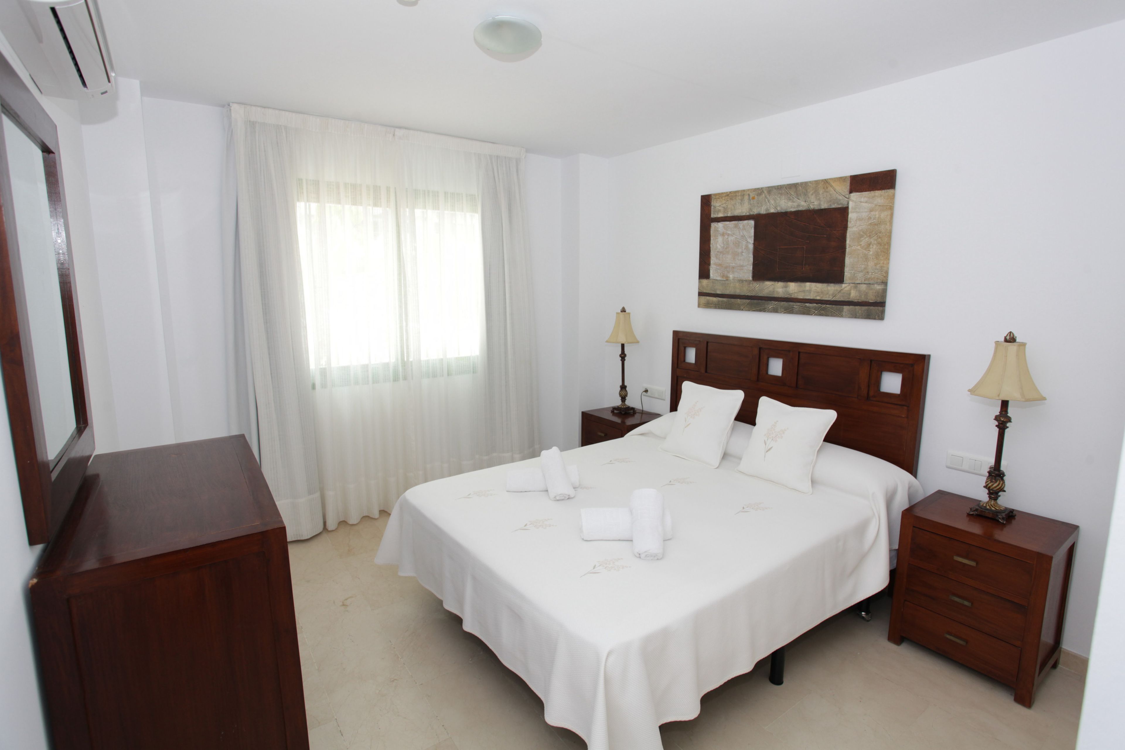 A neatly arranged hotel room with a large bed, two bedside tables with lamps, a wooden headboard, a painting on the wall, and a mirror on the left.