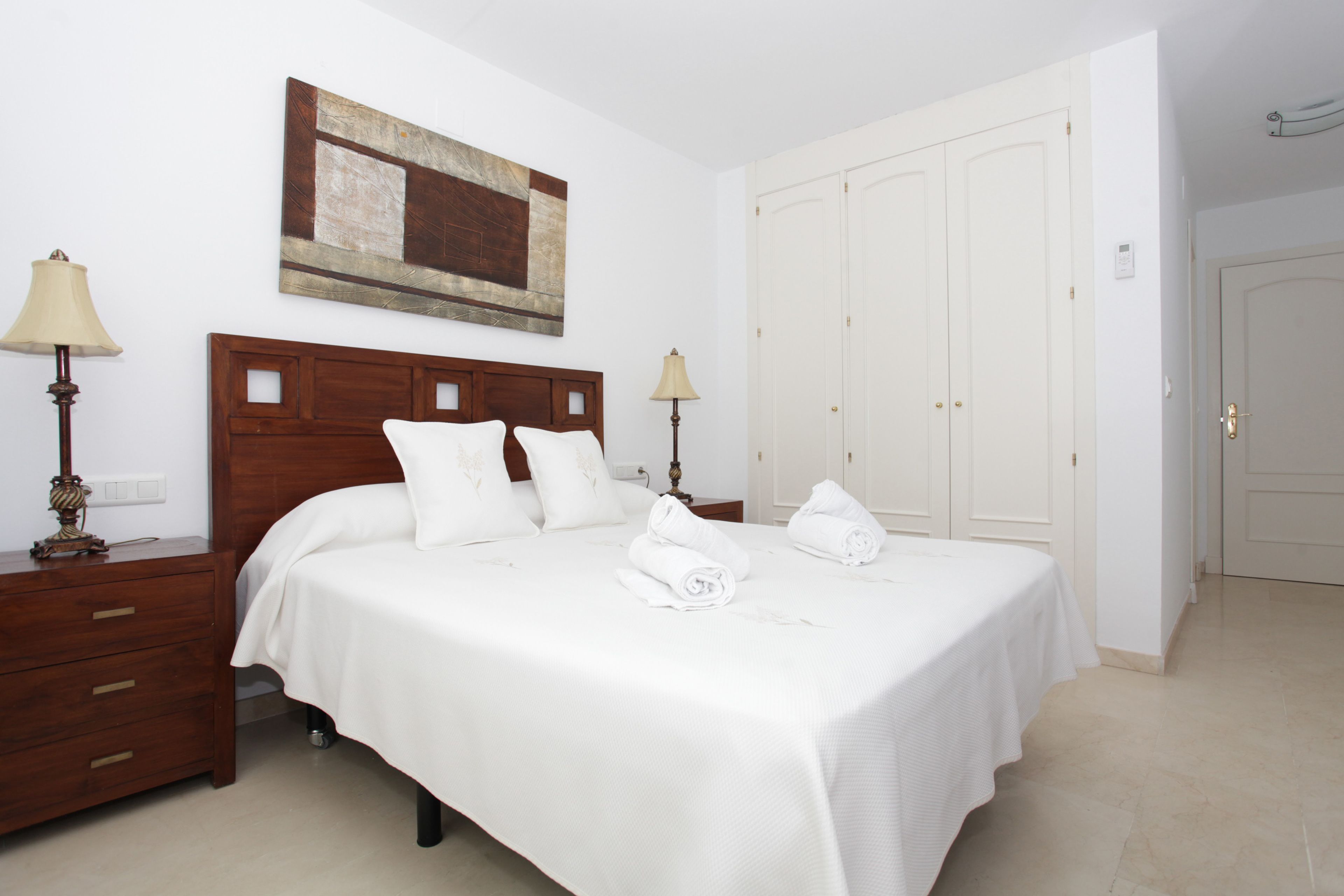 A hotel room with a large bed, two bedside tables with lamps, a wooden headboard, a white door, and a painting on the wall.