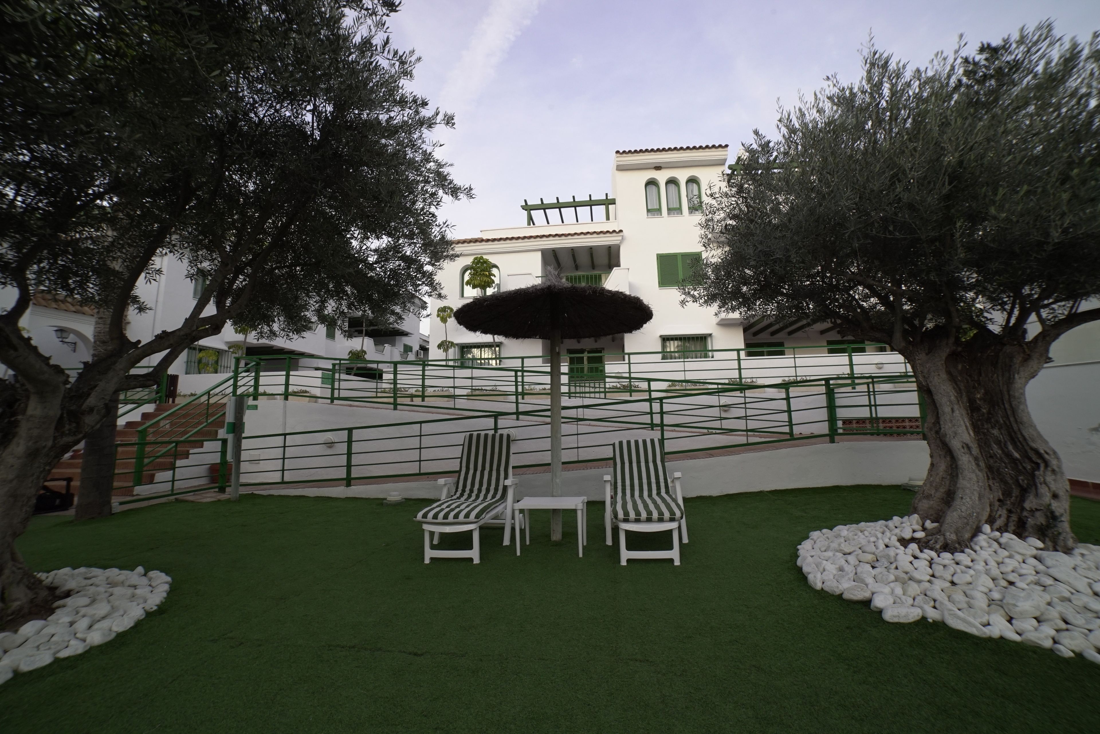 Un patio con dos sillones de descanso, una pequeña mesa y un parasol decorativo. Se ven árboles y un edificio al fondo con fachada blanca y contraventanas verdes.