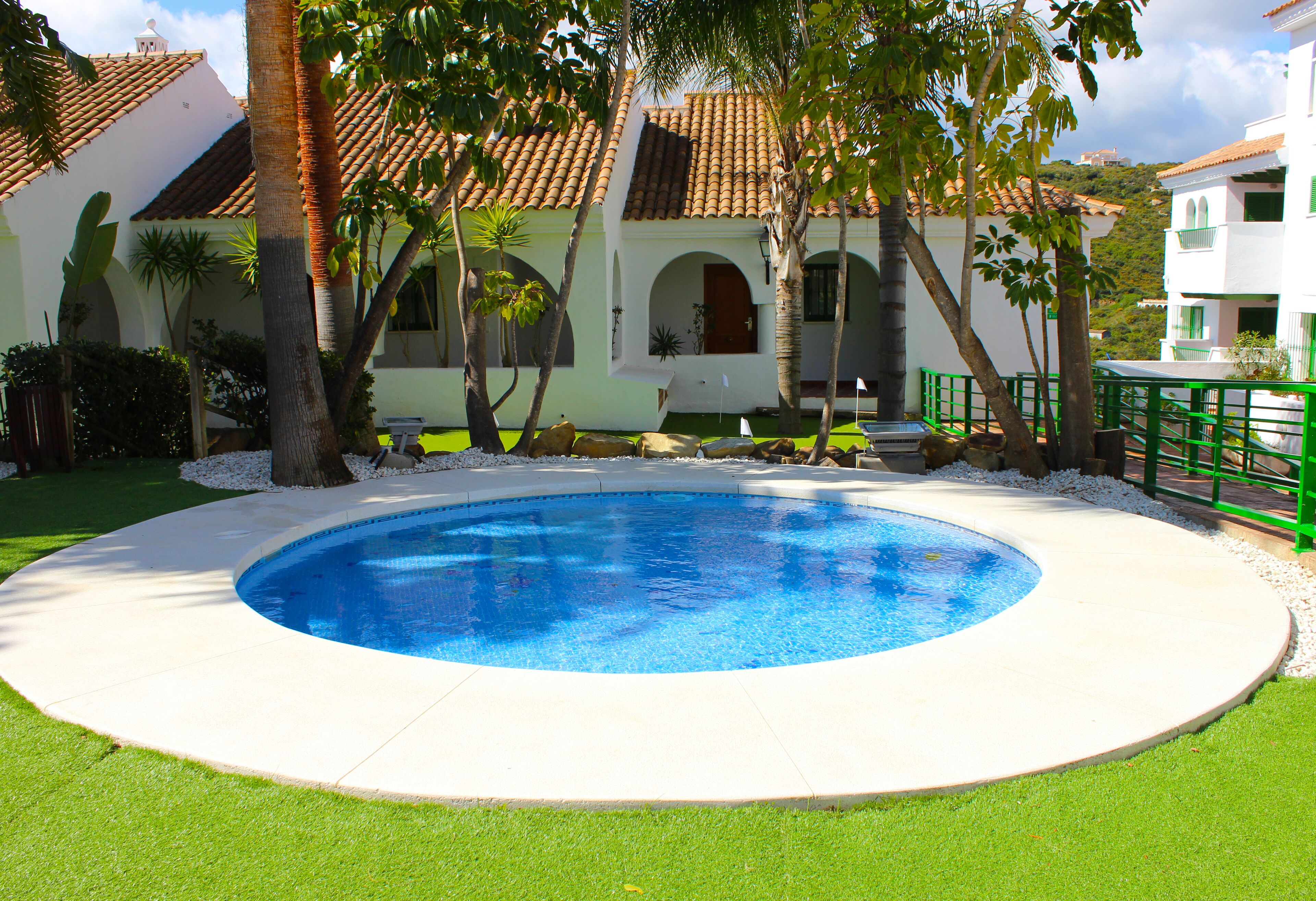 Una piscina rodeada por un deck blanco, con un edificio al fondo que presenta techo de tejas y ventanas arqueadas.