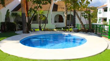 Uma piscina rodeada por um deck branco, com um edifício ao fundo com telhado em azulejos e janelas em arco.