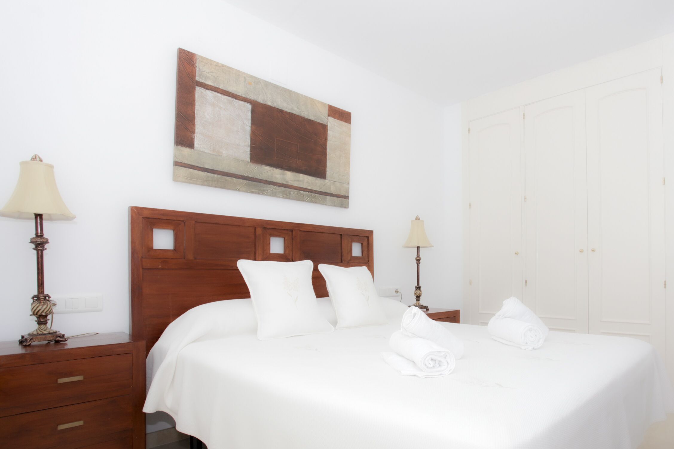 Habitación de hotel con cabecera de madera, ropa de cama blanca, dos lámparas de noche, un cuadro en la pared y un armario.