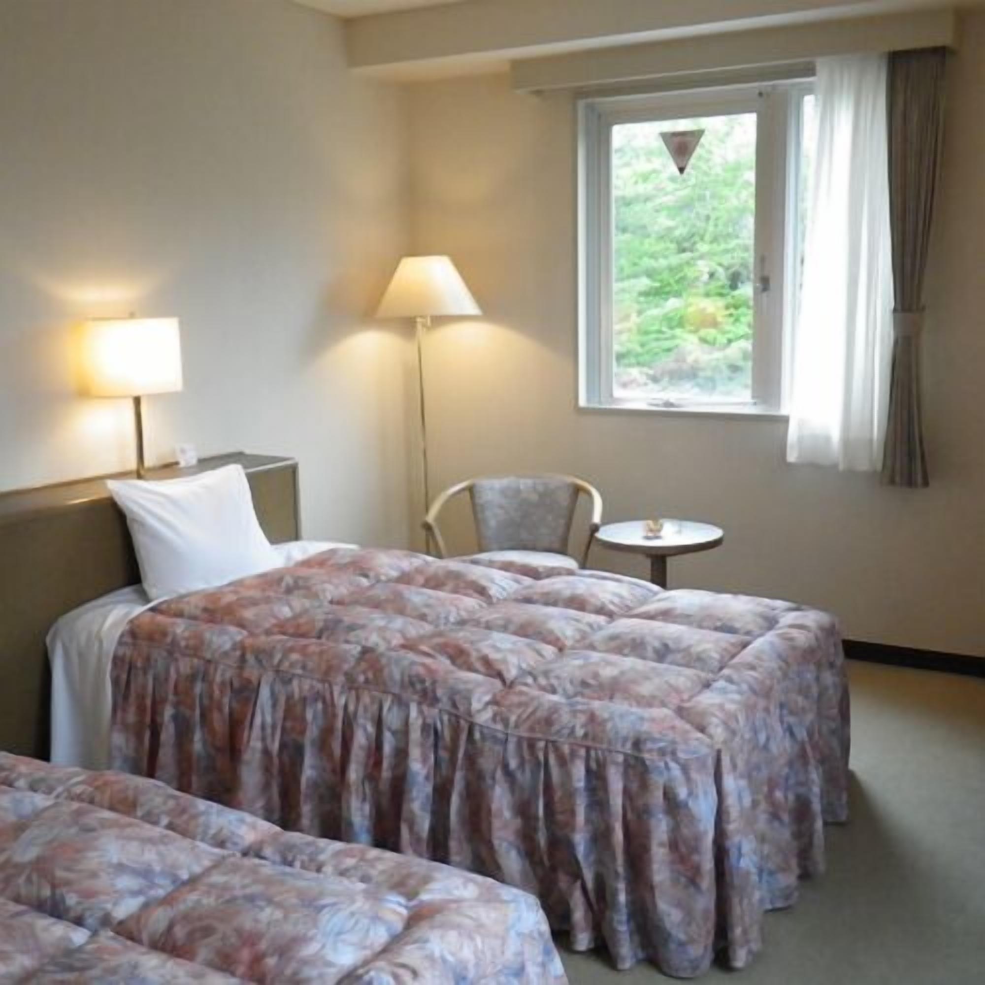 Ein Hotelzimmer mit Bett, zwei Nachttischen, einem Sessel und einem kleinen Tisch, sowie einem Fenster mit Vorhängen.