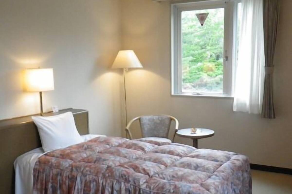 Habitación de hotel con cama, dos mesitas de noche, una silla, una mesita pequeña y una ventana con cortinas.