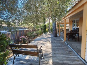 A wooden deck with a bench, a covered patio area with a table and chairs, and a garden with various plants and a fence.