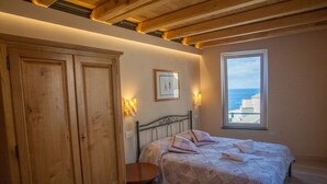 Un dormitorio con cama, armario de madera, una mesita, ventana con vistas al mar y luces fijadas a la pared.