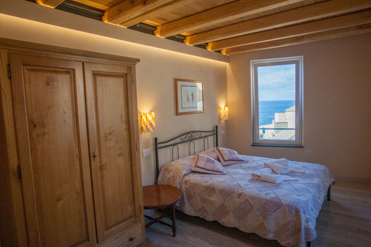 Ein Schlafzimmer mit Bett, Holzschrank, kleinem Tisch, Fenster mit Meerblick und Wandleuchten.