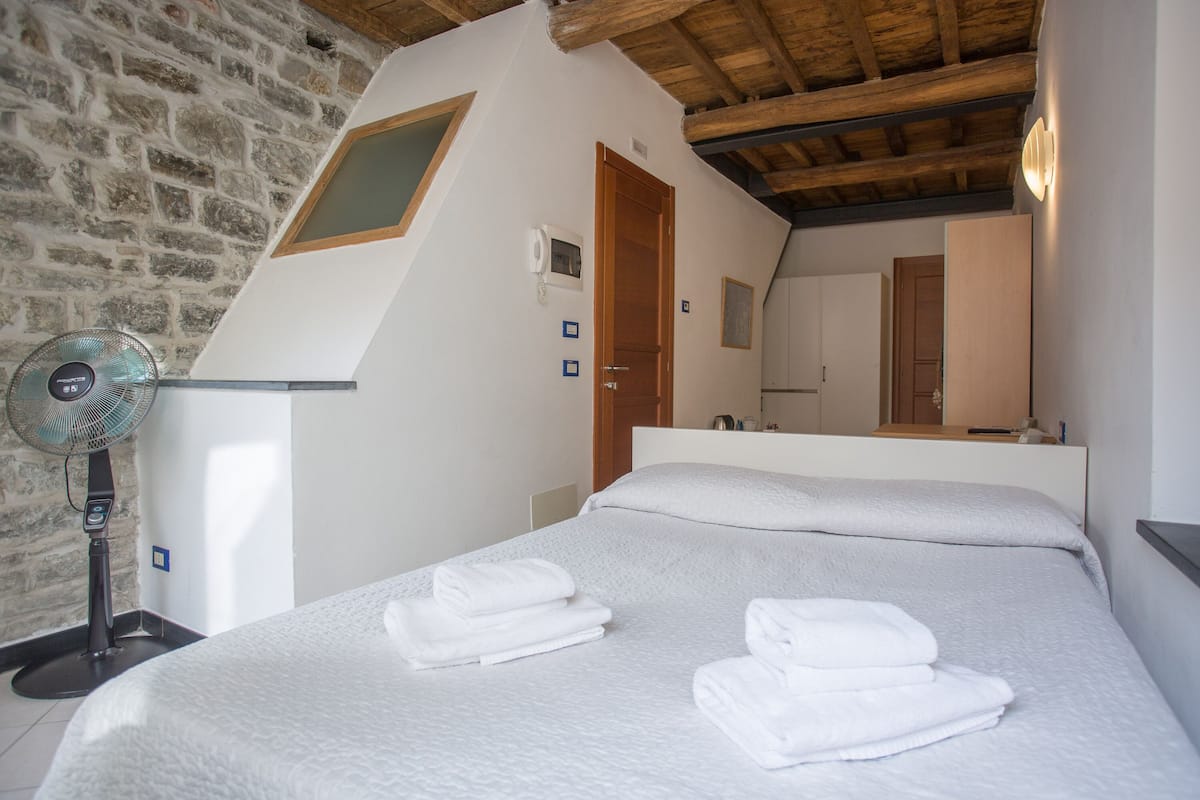 Ein Schlafzimmer mit Steinwand, Holzboden, einem Bett mit weißen Leinen und einem Standventilator sowie einer Holztür.