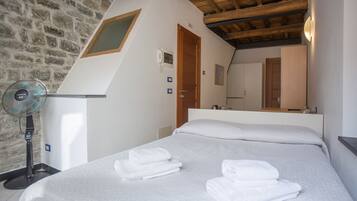 Un dormitorio con pared de piedra, techo de madera, cama con ropa blanca, un ventilador de pie y puerta de madera.