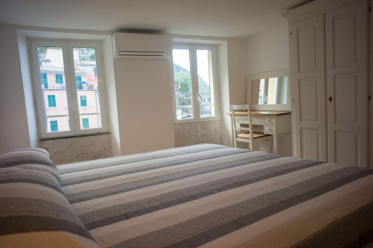 Ein Schlafzimmer mit Bett, Schreibtisch und Kleiderschrank. Fenster bieten Blick auf Gebäude.
