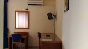 Ein kleines Zimmer mit einem Holztisch, einem Stuhl, einem Fenster mit Jalousien, einer Klimaanlage und einem am Wand montierten Fernseher.