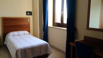 Ein Einzelbett mit Holz-Kopfstück, ein kleiner Schreibtisch, ein Fenster mit Vorhängen und ein an der Wand befestigter Lichtschalter.