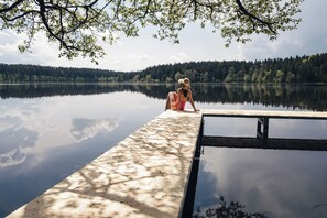 Une femme est assise sur une jetée en bois qui s’avance sur un lac paisible, entourée d’arbres et avec une colline boisée en arrière-plan.