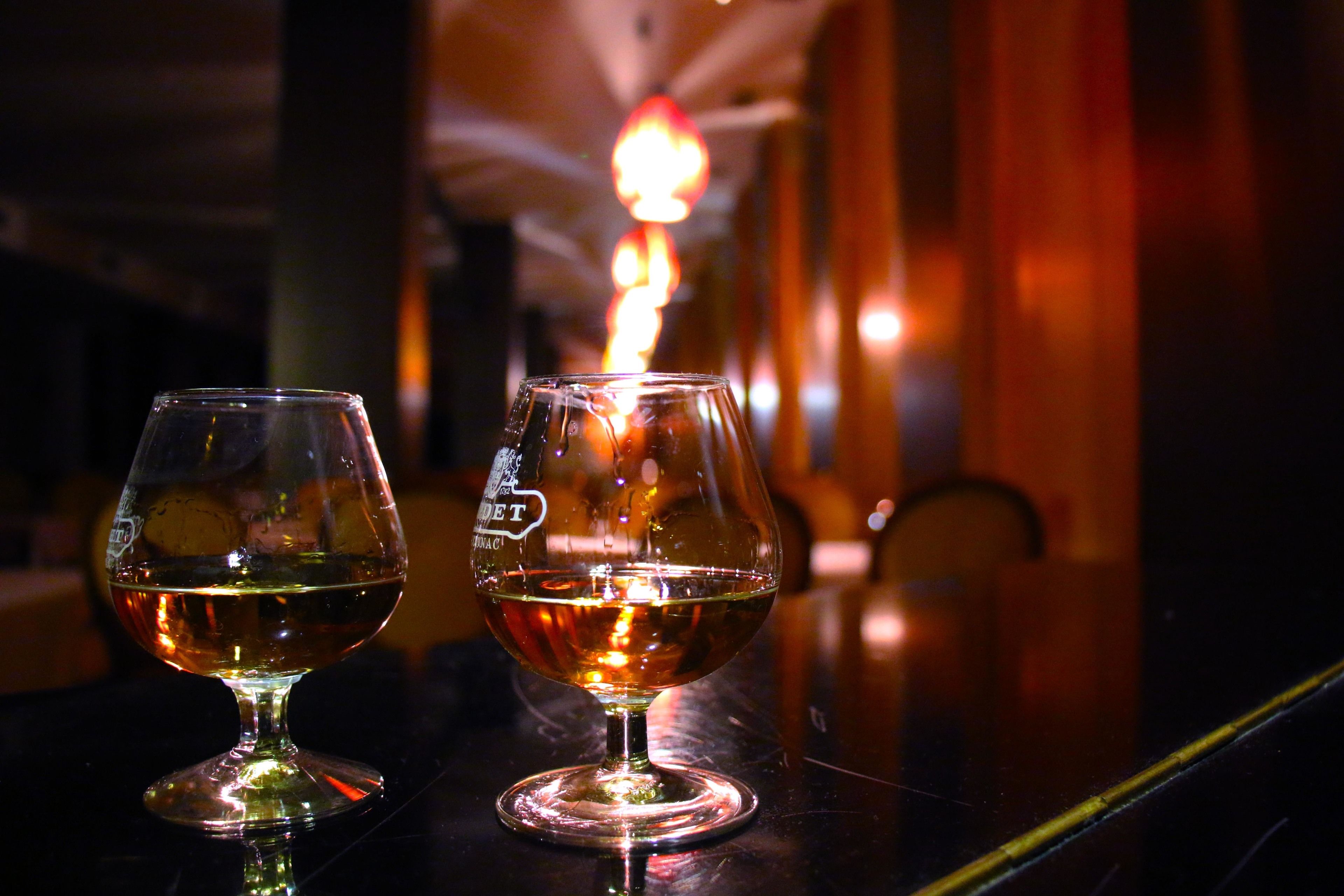 Two glasses of amber liquid on a reflective surface, with a blurred background featuring warm lighting and a vertical element.