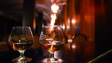 Two glasses of amber liquid on a reflective surface, with a blurred background featuring warm lighting and a vertical element.