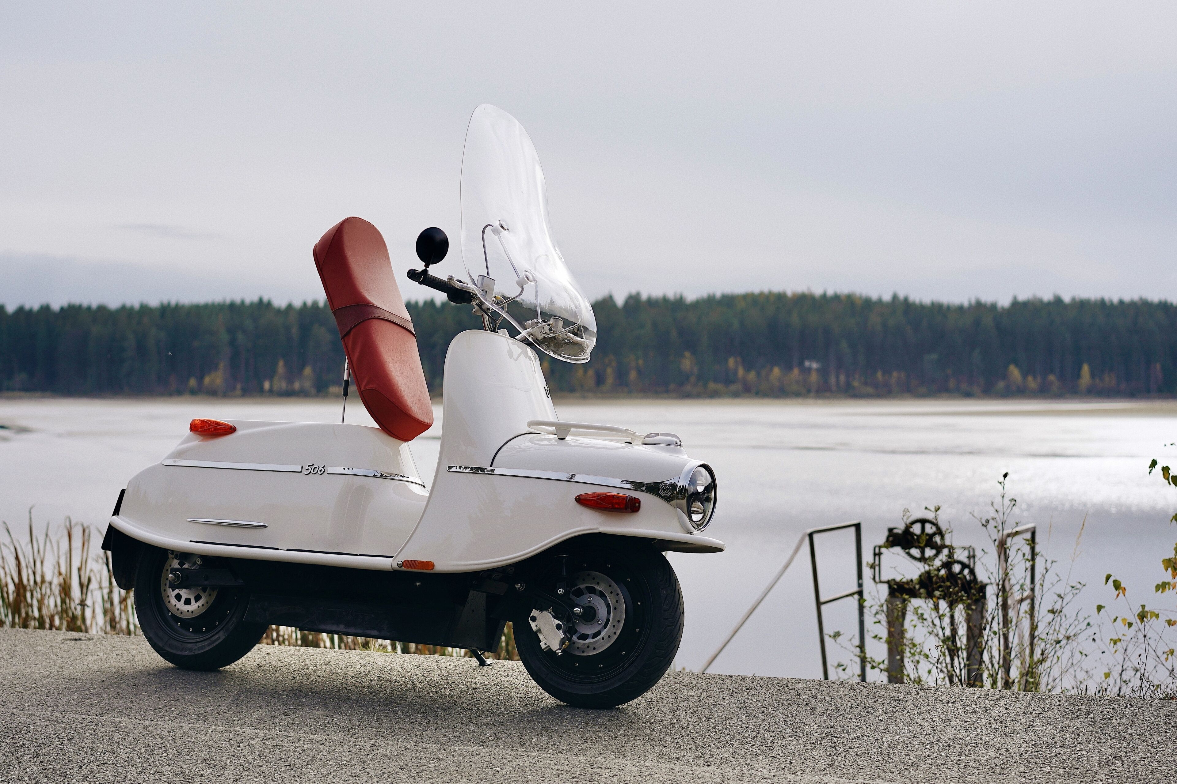 A white scooter with a red seat and a white canopy, parked by a body of water with a forest in the background.