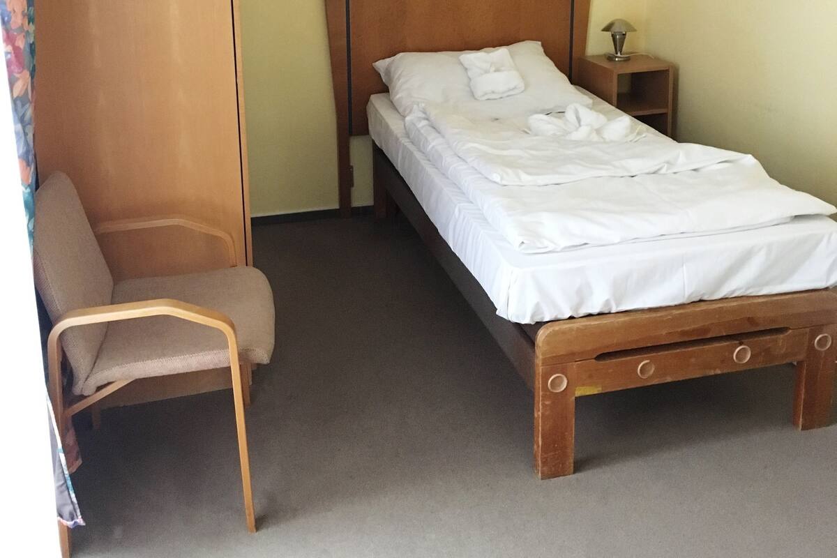 A bedroom with a wooden bed, a chair, a nightstand, and a wall-mounted light fixture.