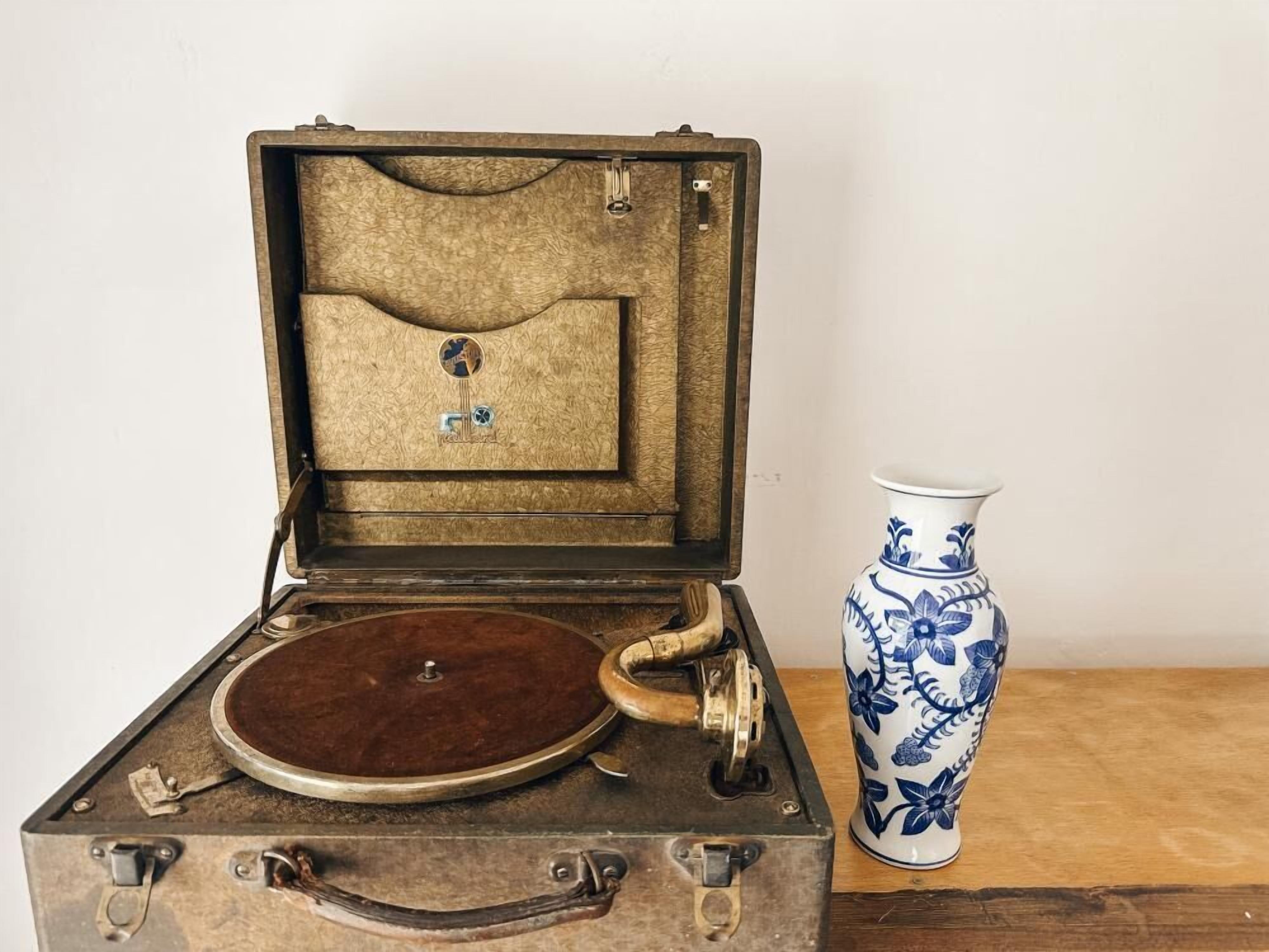 An open vintage suitcase with a record player inside, placed next to a blue and white floral vase on a wooden surface.