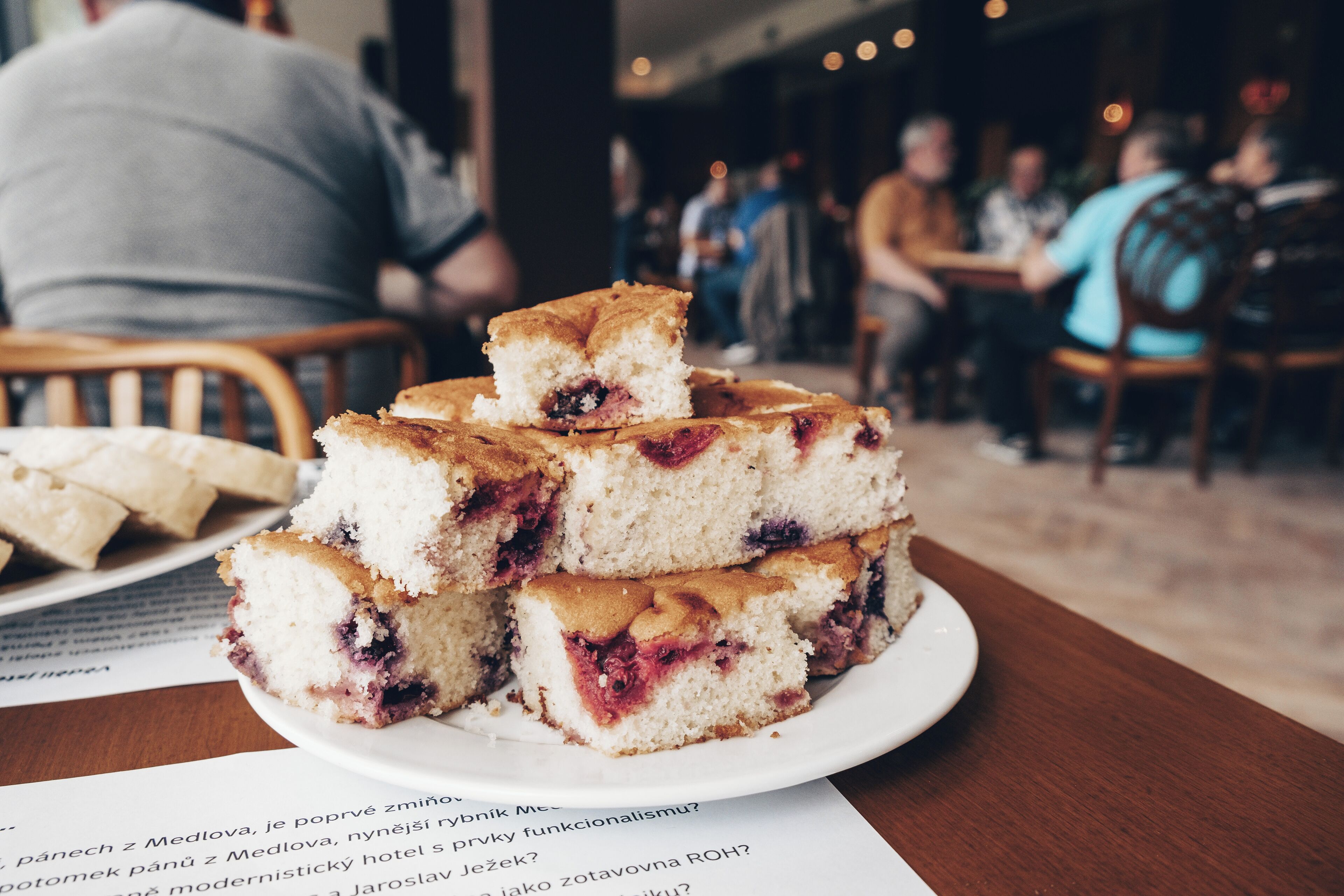 A stack of layered cakes on a white plate, with a blurred background of people dining.