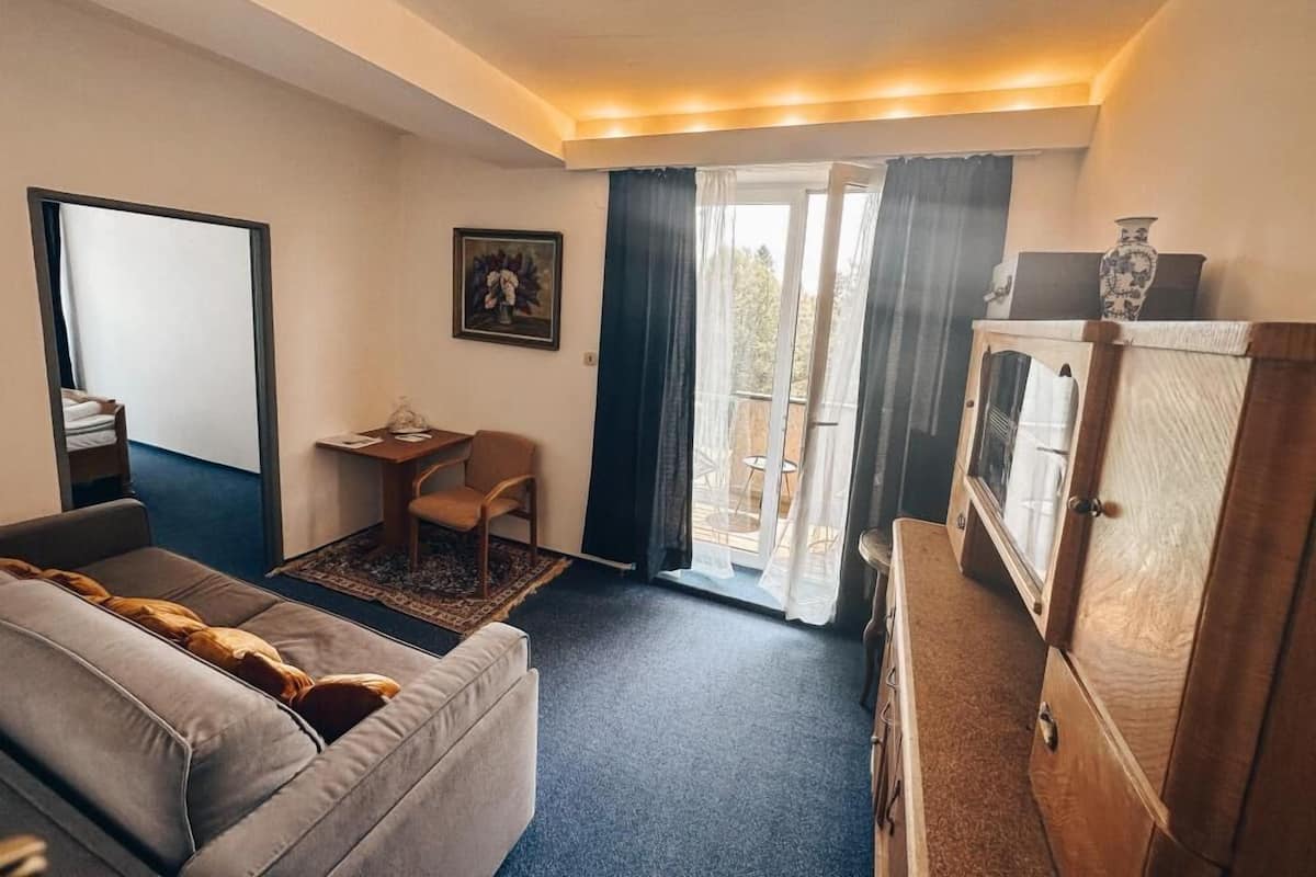 A hotel room with a sofa, a small table with a chair, a cabinet, and a view of the outdoors through a glass door.