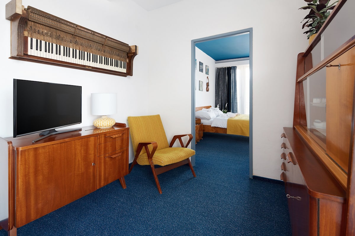 A hotel room with a large flat-screen TV, a wooden cabinet, a yellow armchair, a bed with a blue headboard, and a wooden wardrobe.