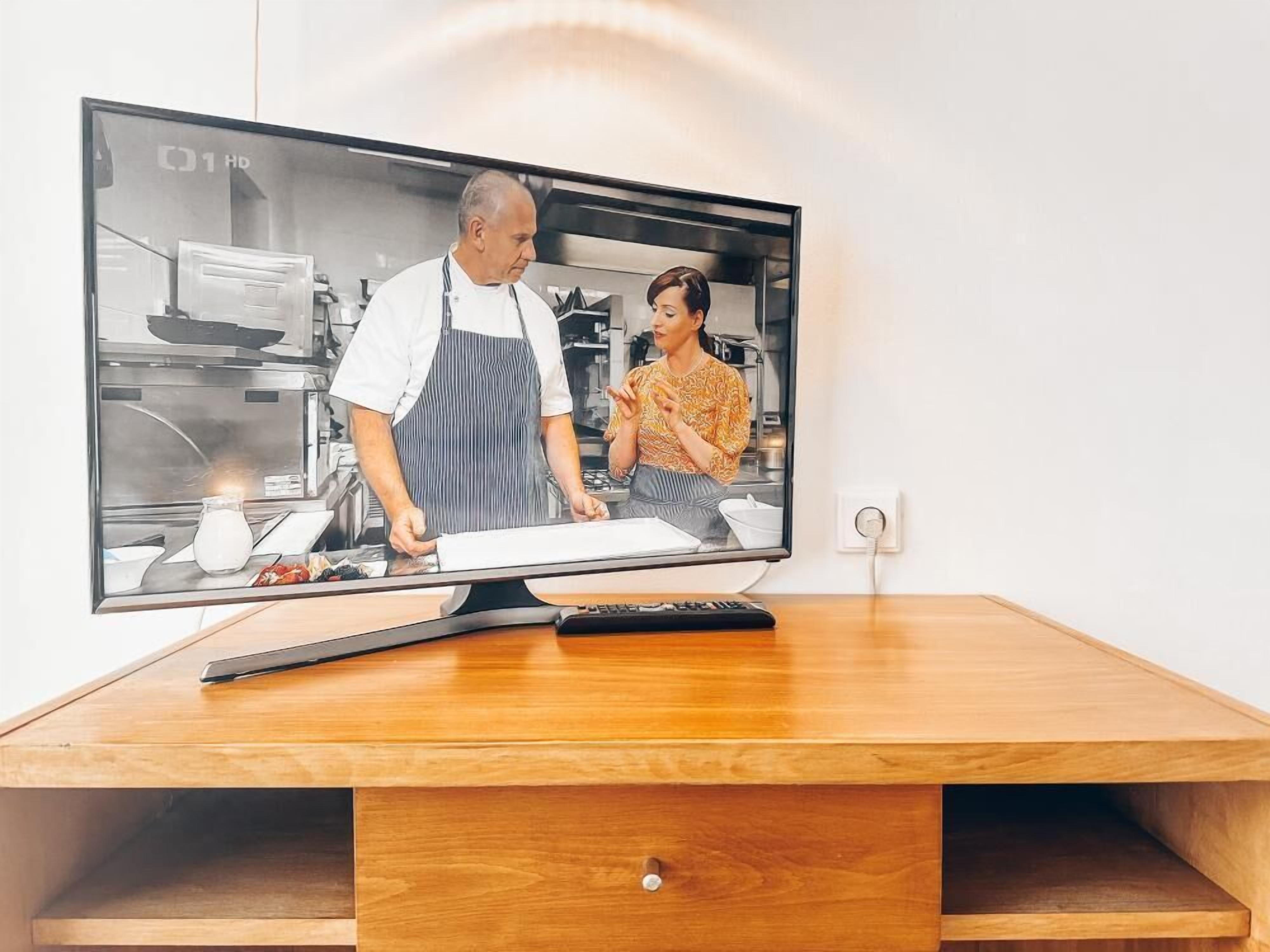 A television mounted on a wooden desk with a remote control, displaying a kitchen scene.