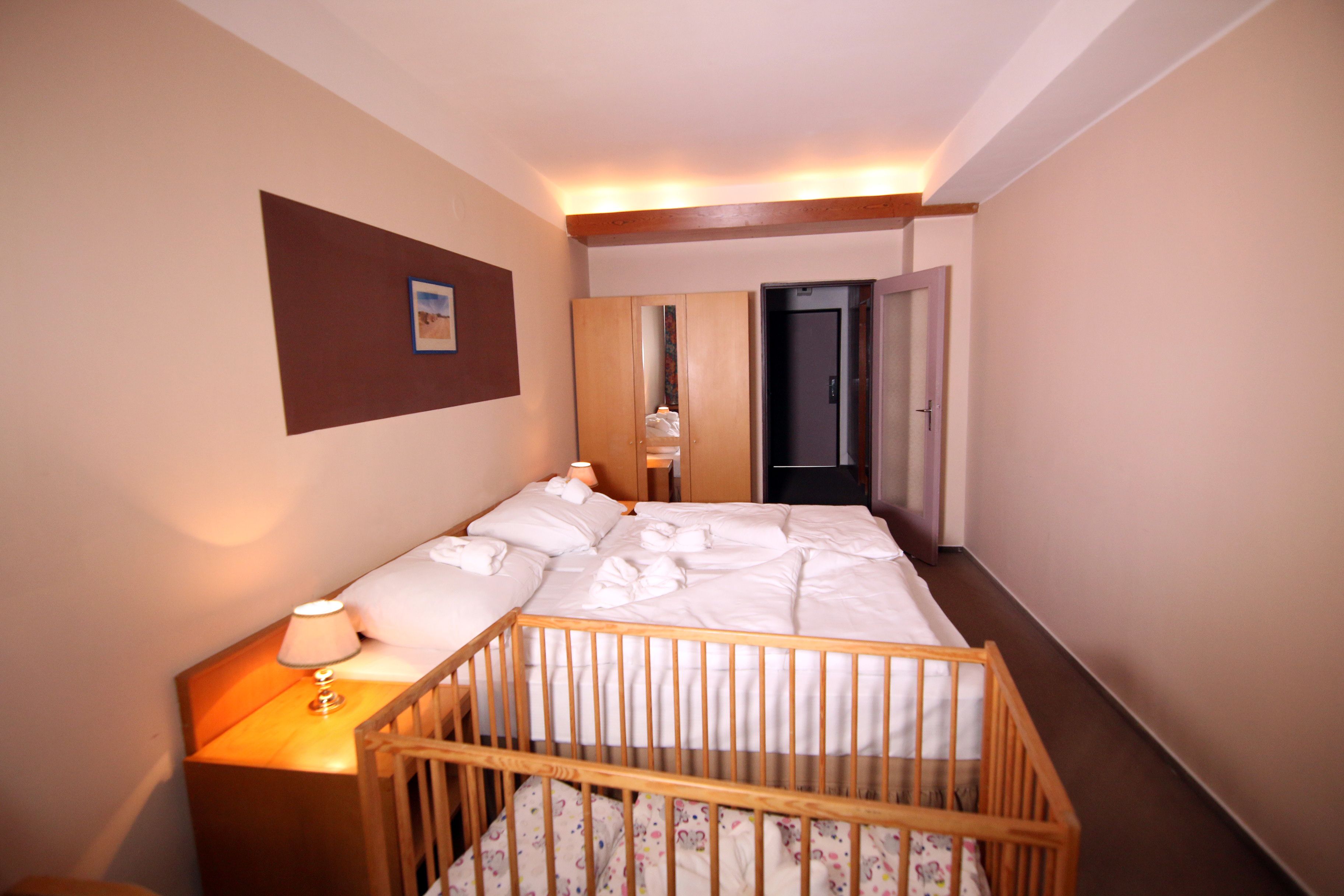 A hotel room with a wooden crib, a bed with white linens, a nightstand with a lamp, a mirror, and a door leading to another room.